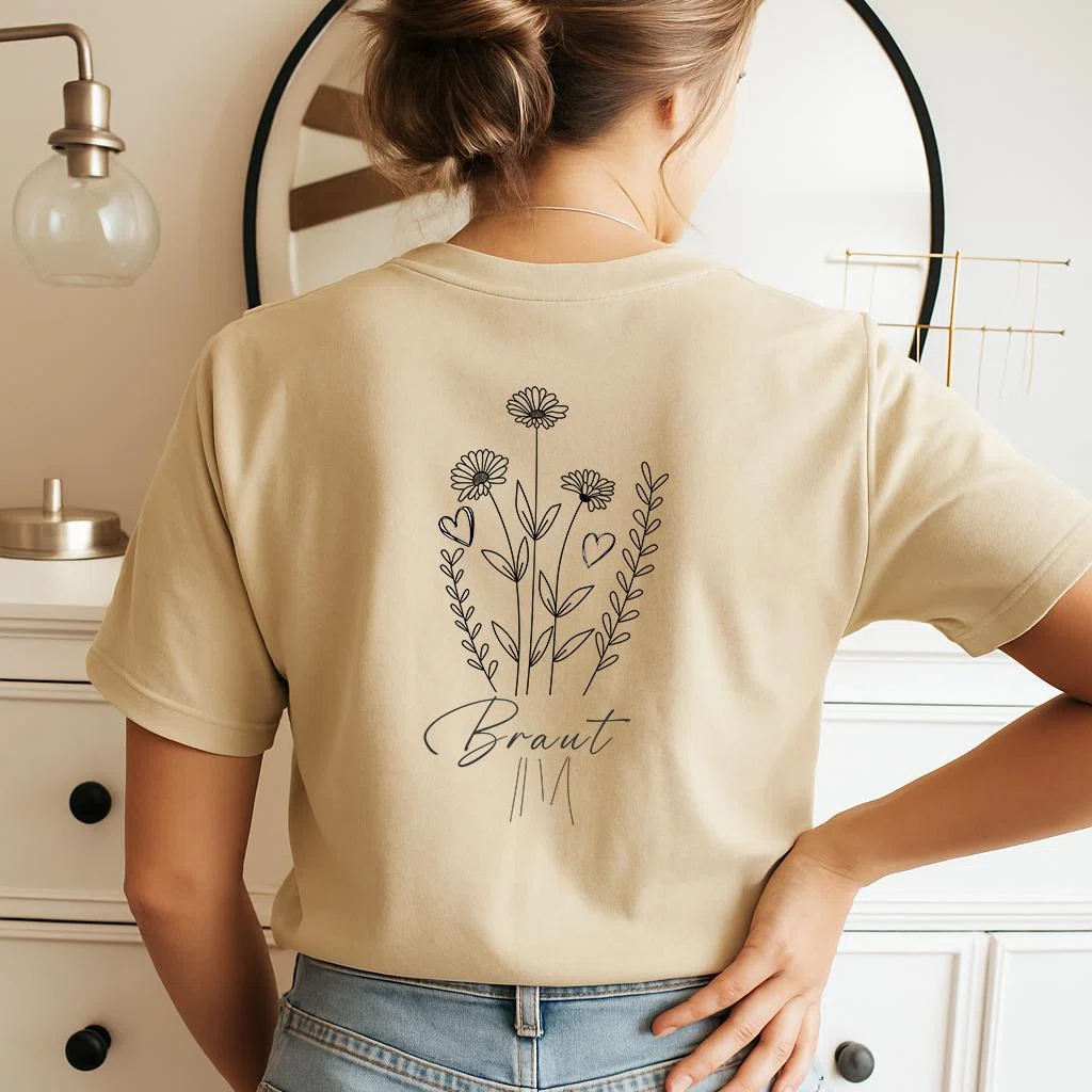 Beiges Kurzarmshirt in Rückansicht mit schwarzer Blumenzeichnung aus Margeriten, Blätterranken und kleinen Herzen; darunter steht der Schriftzug „Braut“.