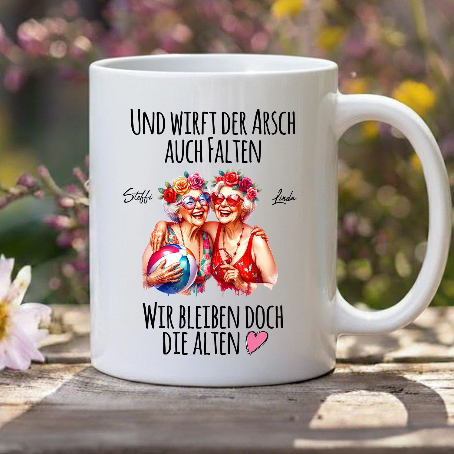 Weiße Tasse, frontal mit Henkel rechts, bedruckt mit zwei illustrierten Frauen in Rot, Sonnenbrillen, Blumenkränzen und Wasserball. Dazu der Spruch „Und wirft der Arsch auch Falten – wir bleiben doch die Alten“ mit Herz.