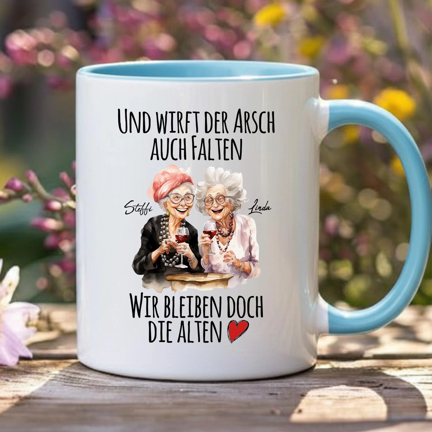 Weiße Tasse mit hellblauer Innenseite und Henkel, frontal fotografiert. Der Aufdruck zeigt zwei gezeichnete ältere Damen mit Weingläsern, ein rotes Herz und den Spruch „Und wirft der Arsch auch Falten, wir bleiben doch die Alten“.