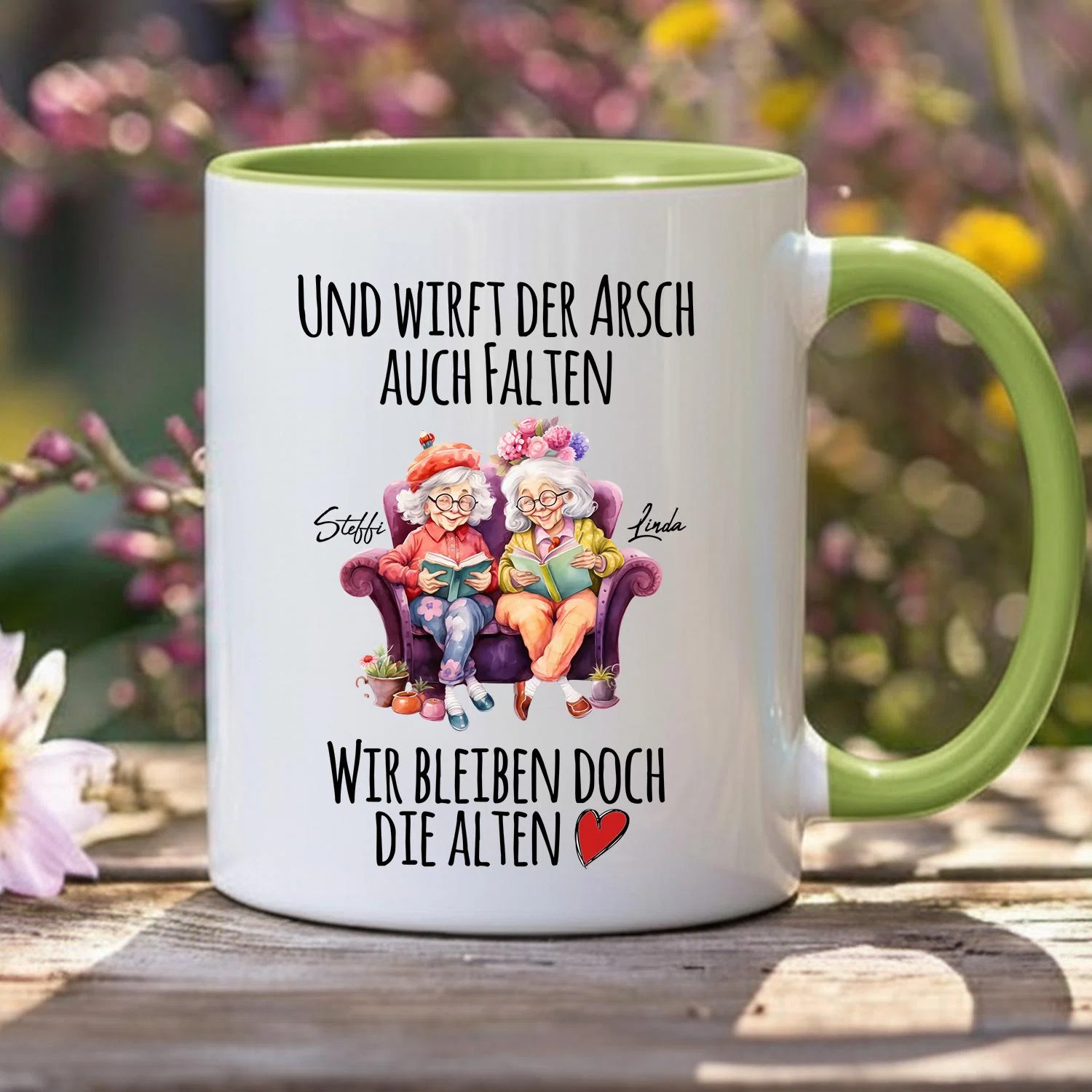 Weiße Tasse mit hellgrünem Henkel und Innenrand, frontal auf einem Holztisch. Aufdruck mit zwei älteren Frauen beim Lesen auf einem violetten Sofa und dem Spruch „Und wirft der Arsch auch Falten, wir bleiben doch die Alten“ samt rotem Herz.