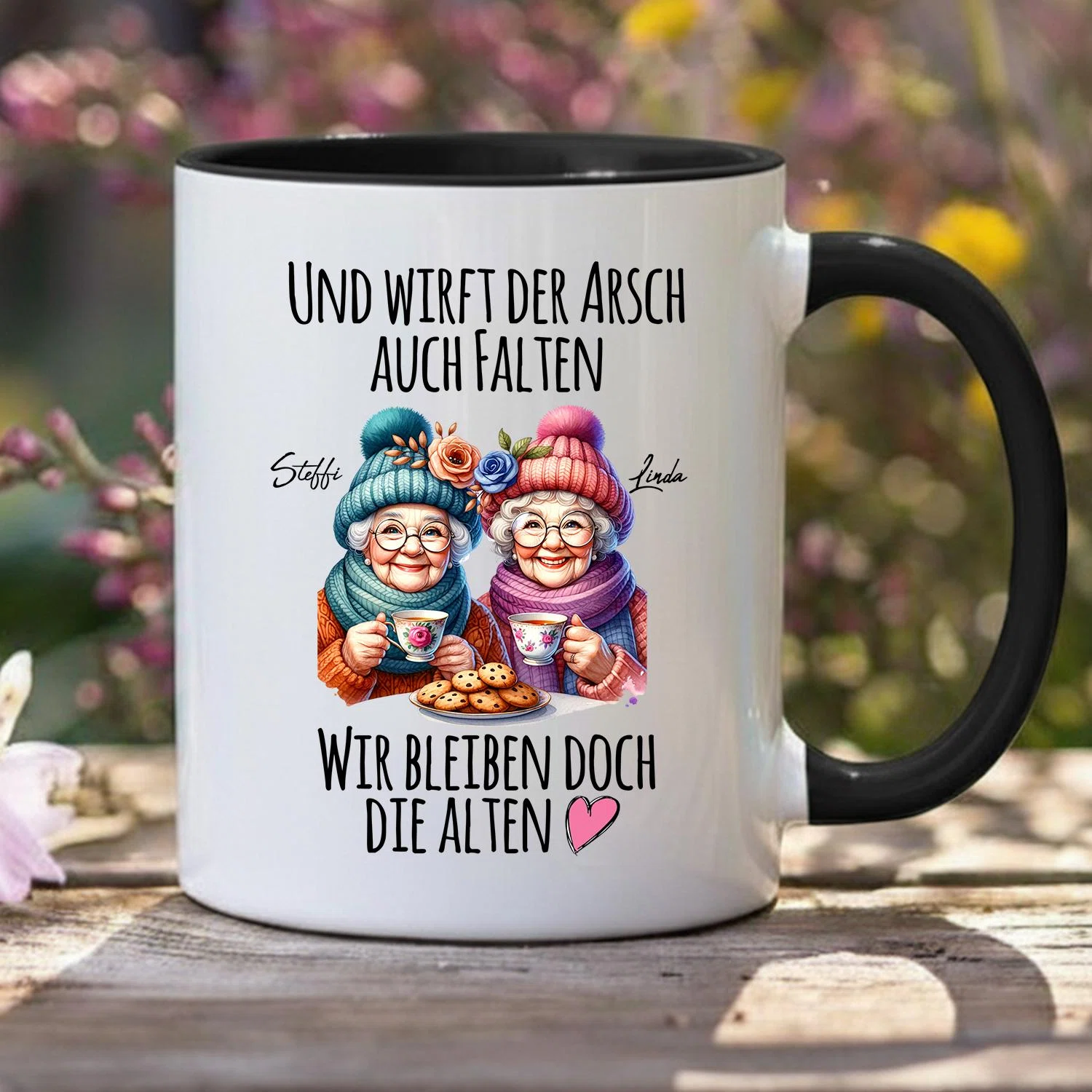 Weiße Tasse mit schwarzem Henkel und schwarzer Innenseite, frontal fotografiert. Zwei illustrierte ältere Frauen mit bunten Mützen halten Teetassen über einem Teller Kekse; dazu der Spruch „Und wirft der Arsch auch Falten – wir bleiben doch die Alten“.