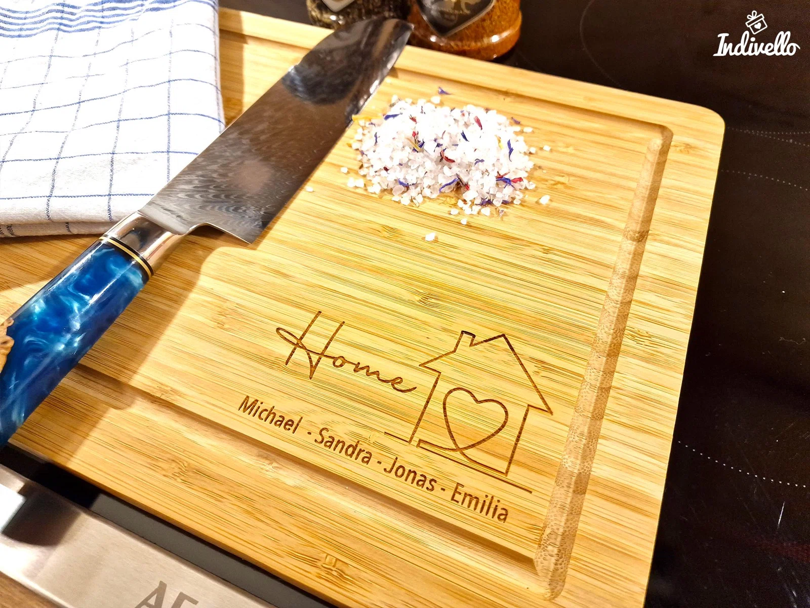 Helles rechteckiges Schneidebrett mit Saftrille, personalisierter Namensgravur und „Home“-Motiv aus Hausumriss und Herz. Schräg von oben mit Messer und grobem Salz inszeniert.