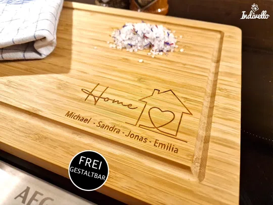 Schneidebrett mit Gravur bestellen: hellbraunes Brett mit umlaufender Saftrille, „Home“-Schriftzug, Haus mit Herz und persönlicher Namenszeile, schräg von oben fotografiert.