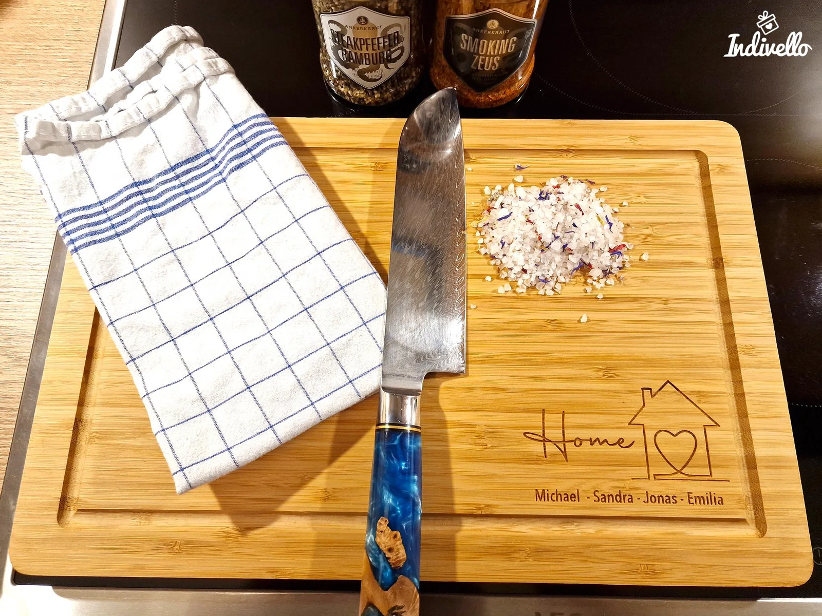 Rechteckiges Schneidebrett in heller Holzoptik mit umlaufender Saftrille und Gravur aus „Home“, Haus mit Herz sowie vier Beispielnamen; schräg von oben mit Messer, Tuch und Gewürzsalz fotografiert.