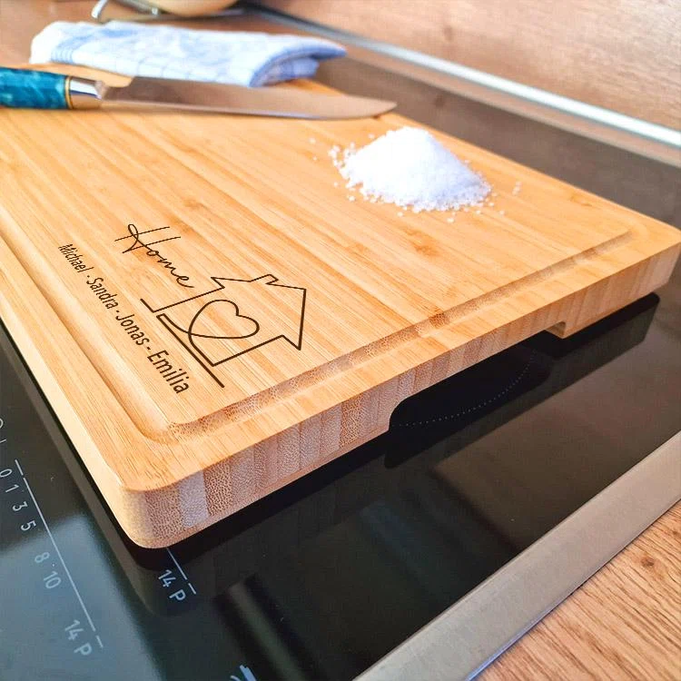 Rechteckiges Schneidebrett in heller Holzoptik mit Saftrille und personalisierter Gravur: Hausumriss mit Herz, „Home“ und mehrere Namen. Schräg auf einem Kochfeld neben Messer und Salz platziert.