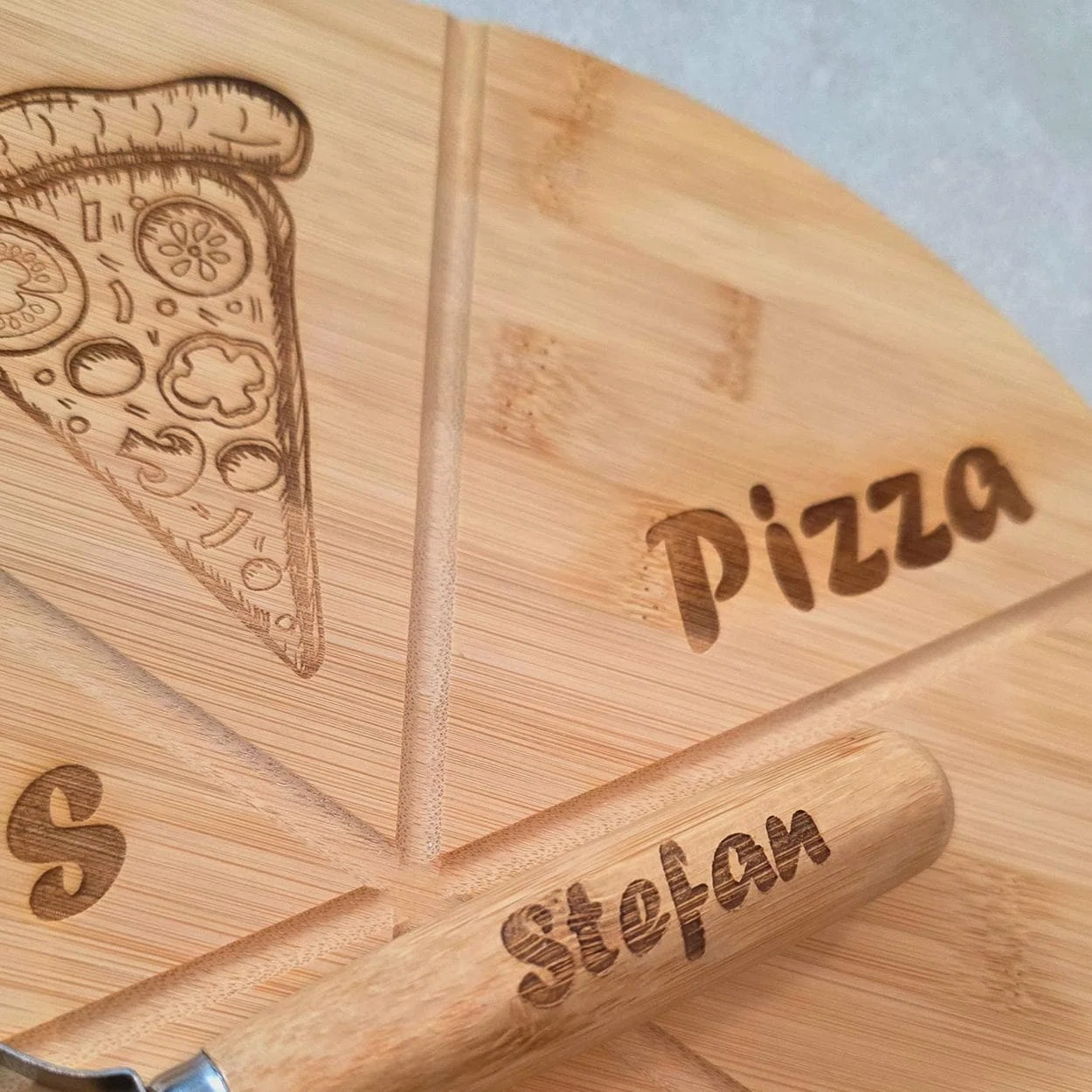 Nahaufnahme eines runden, hellbraunen Pizzabretts mit eingraviertem Pizzastück, Schriftzug „Pizza“ und Rillen zur Stückeinteilung. Darauf liegt ein Pizzaschneider mit graviertem Beispielnamen.