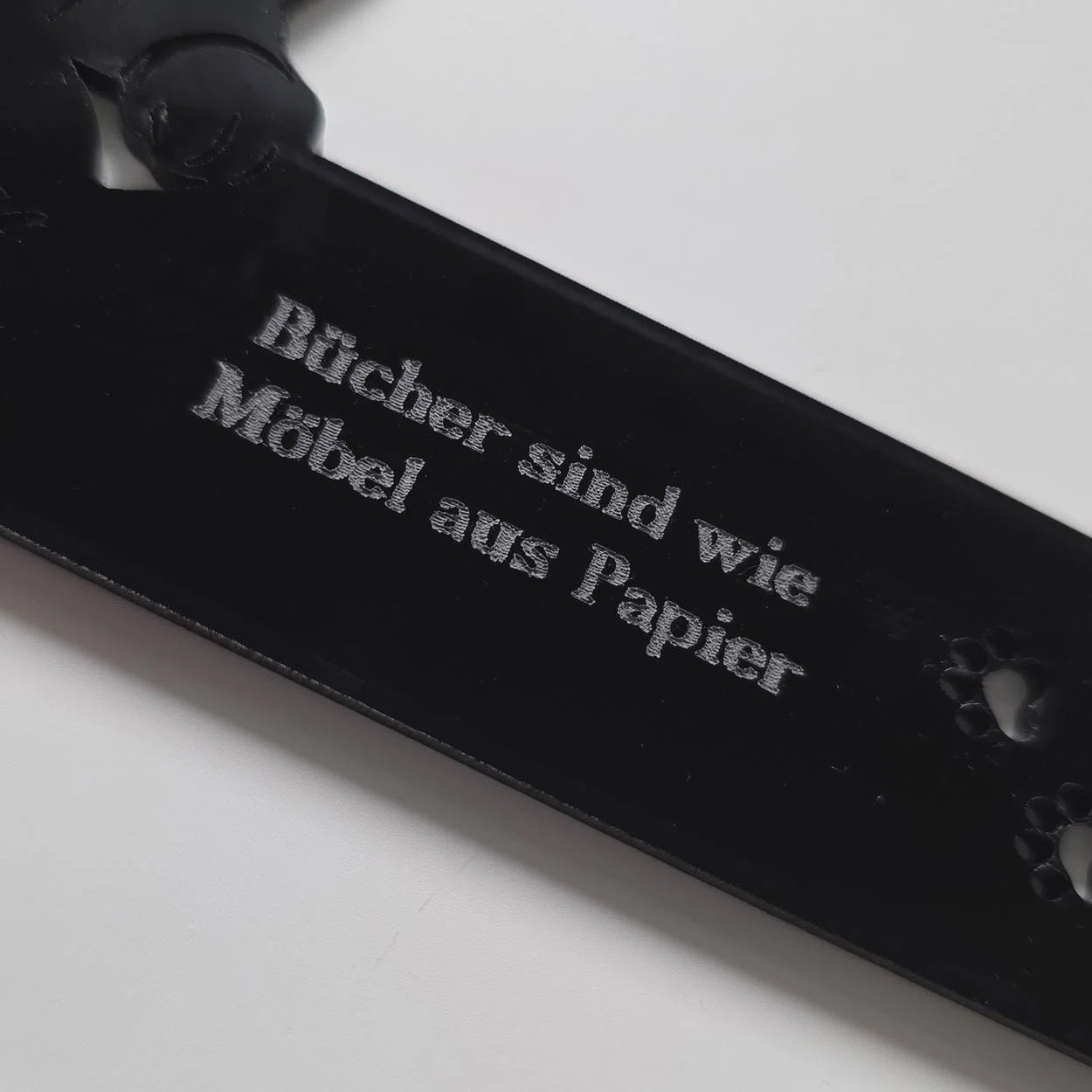 Schwarzer, länglicher Anhänger mit silberfarbener Aufschrift „Bücher sind wie Möbel aus Papier“ und kleinen Pfotenmotiven, diagonal in Nahaufnahme fotografiert.