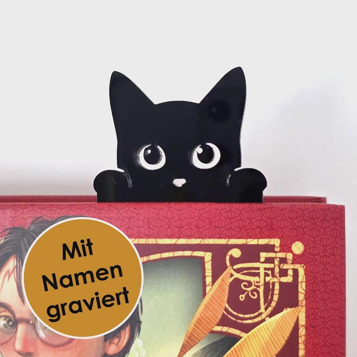 Schwarzes Lesezeichen in Katzenform, das mit großen weißen Augen und zwei Pfoten über den Rand eines roten Buches schaut. Ein Hinweis nennt die Gravur mit Namen.