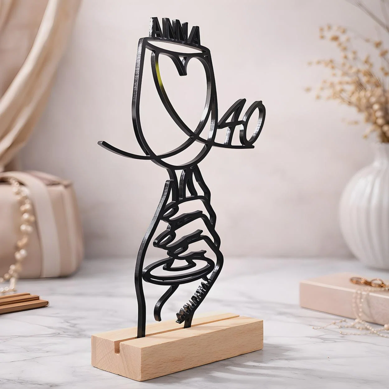 Schwarze, personalisierte Linienfigur einer Hand, die einen herzförmigen Ballon mit zwei Namen hält, frontal in einem hellen rechteckigen Standfuß präsentiert.