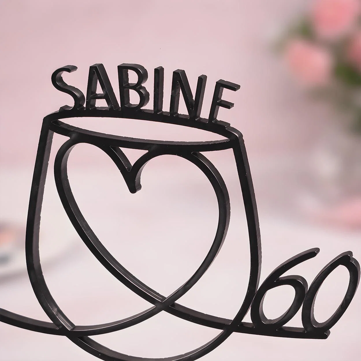 Schwarzer, filigraner Deko-Topper in Linienoptik mit großem Herz, dem Schriftzug „SABINE“ und der Zahl 60, frontal vor rosafarbenem Hintergrund fotografiert.