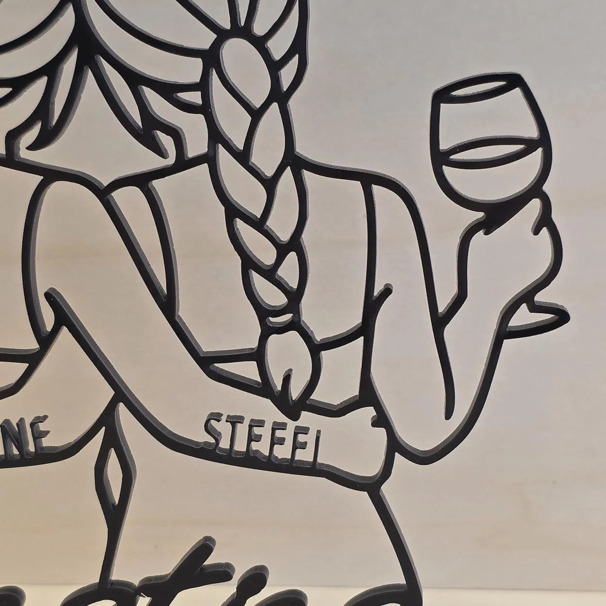 Nahaufnahme eines schwarzen Kontur-Aufstellers vor hellem Hintergrund: Eine Figur mit langem Zopf hält ein Weinglas; auf dem schrägen Band steht der ausgeschnittene Name „STEFFI“.