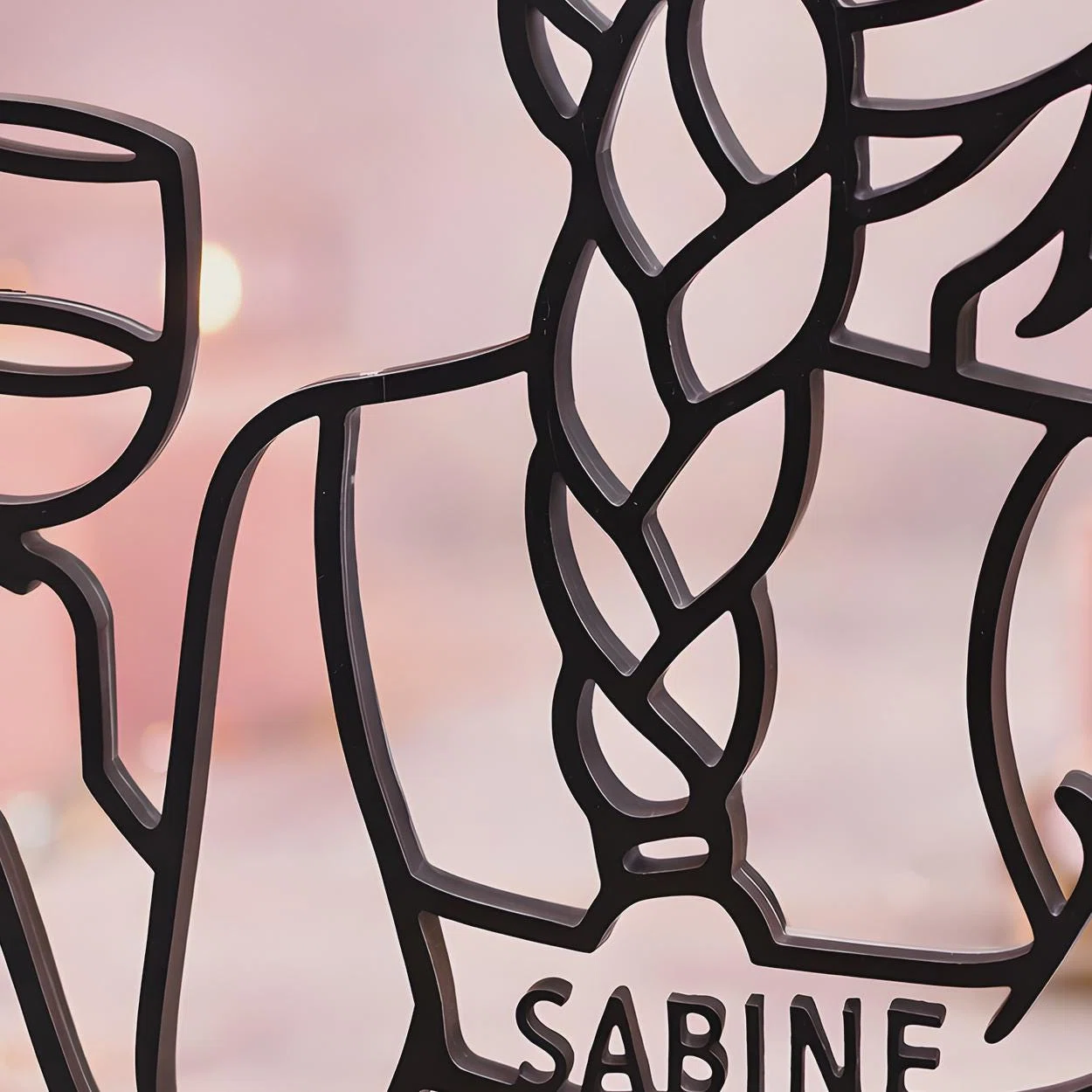 Nahaufnahme eines schwarzen, personalisierten Deko-Aufstellers im Linienlook mit stilisierten Gesichtern, Brille und geflochtenem Haar vor weich verschwommenem rosa Hintergrund.