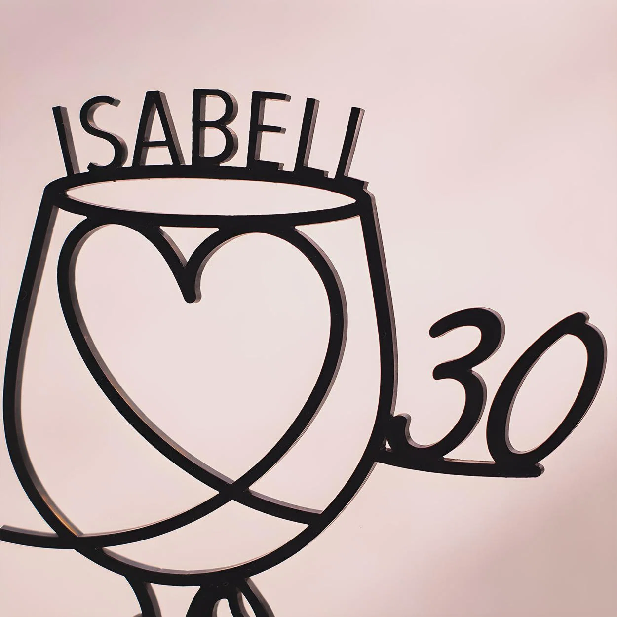 Schwarzer, personalisierter Tortenstecker in Form eines Weinglases mit großem Herz in der Mitte, dem Namen „Isabell“ am Rand und der Zahl 30 seitlich, frontal vor rosafarbenem Hintergrund.