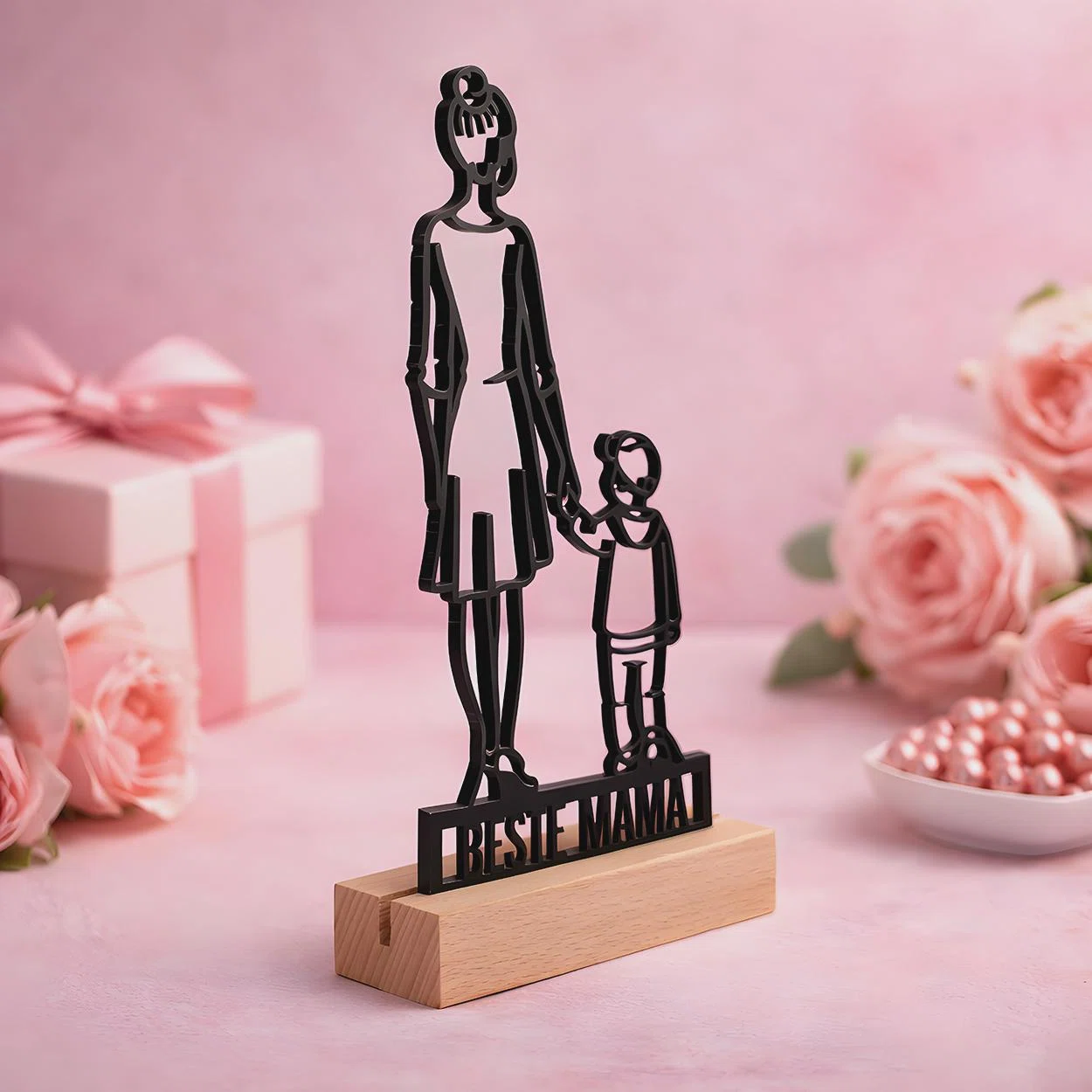 Schwarze Aufstellfigur mit dem Motiv einer Frau, die ein Kind an der Hand hält, über dem Schriftzug „BESTE MAMA“. In einem hellen Sockel vor rosa Kulisse mit Rosen und Geschenk präsentiert.