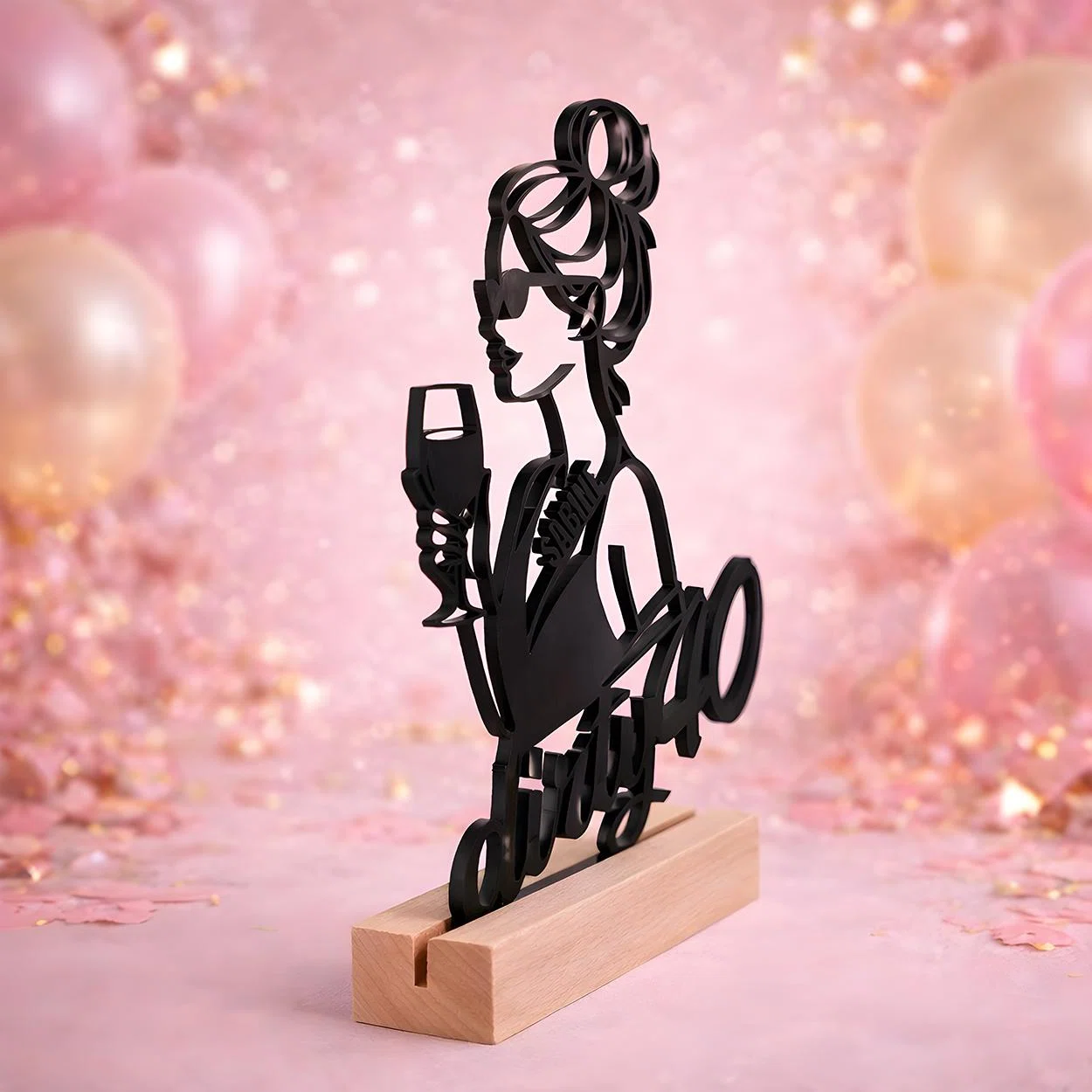 Schwarzes Dekoschild als stilisierte Silhouette einer Frau mit Hochsteckfrisur, Sonnenbrille und Sektglas, frontal in einem hellen Sockel vor rosa glitzernder Ballonkulisse präsentiert.