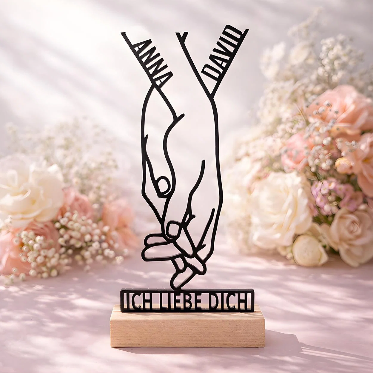Schwarze Linienfigur zweier Hände mit verschränkten kleinen Fingern und dem Schriftzug „ICH LIEBE DICH“, frontal auf einem hellen, holzfarbenen Sockel vor rosa-weißen Blumen arrangiert.