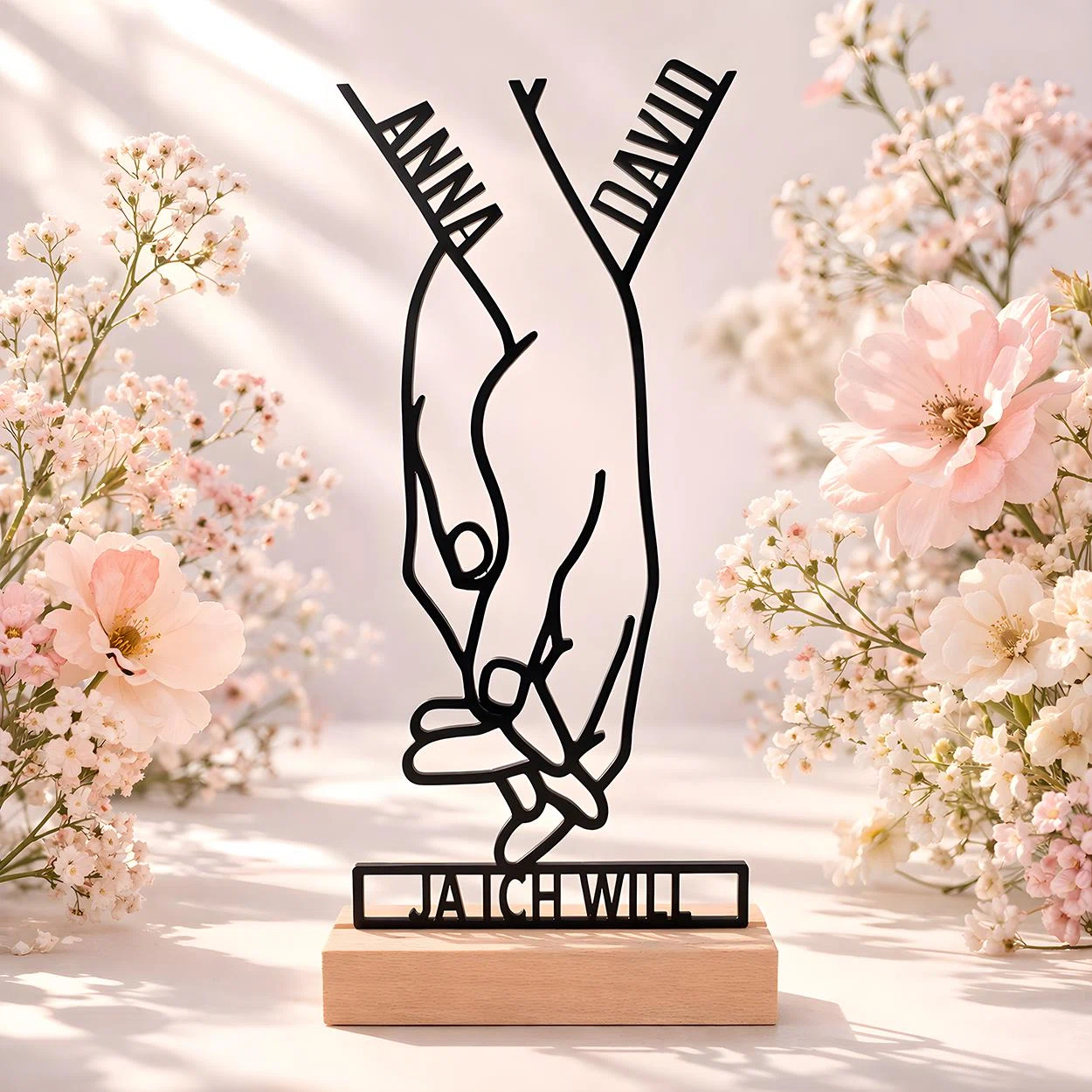 Schwarze Dekofigur in feiner Linienoptik mit zwei ineinandergreifenden Händen und Ringmotiv. Der Schriftzug „JA ICH WILL“ steht auf einem hellen Sockel, umrahmt von rosa Blüten.