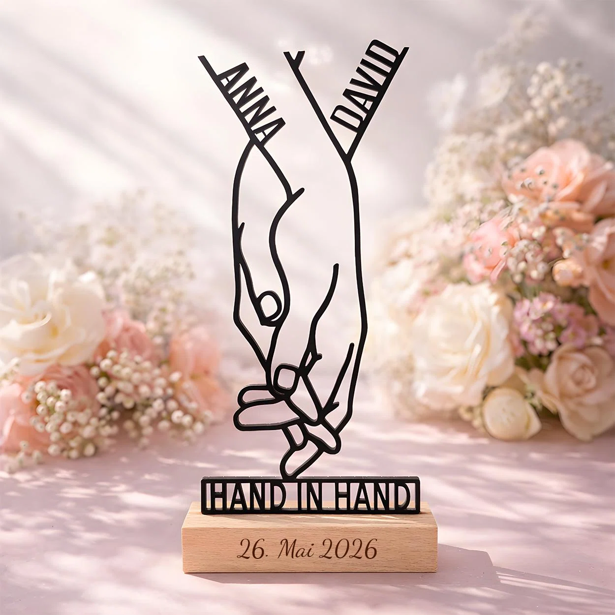 Schwarze Linienfigur zweier Hände mit ineinander verhakten kleinen Fingern und Schriftzug „HAND IN HAND“ auf hellbraunem Sockel mit graviertem Datum, frontal vor rosa-weißen Blüten.