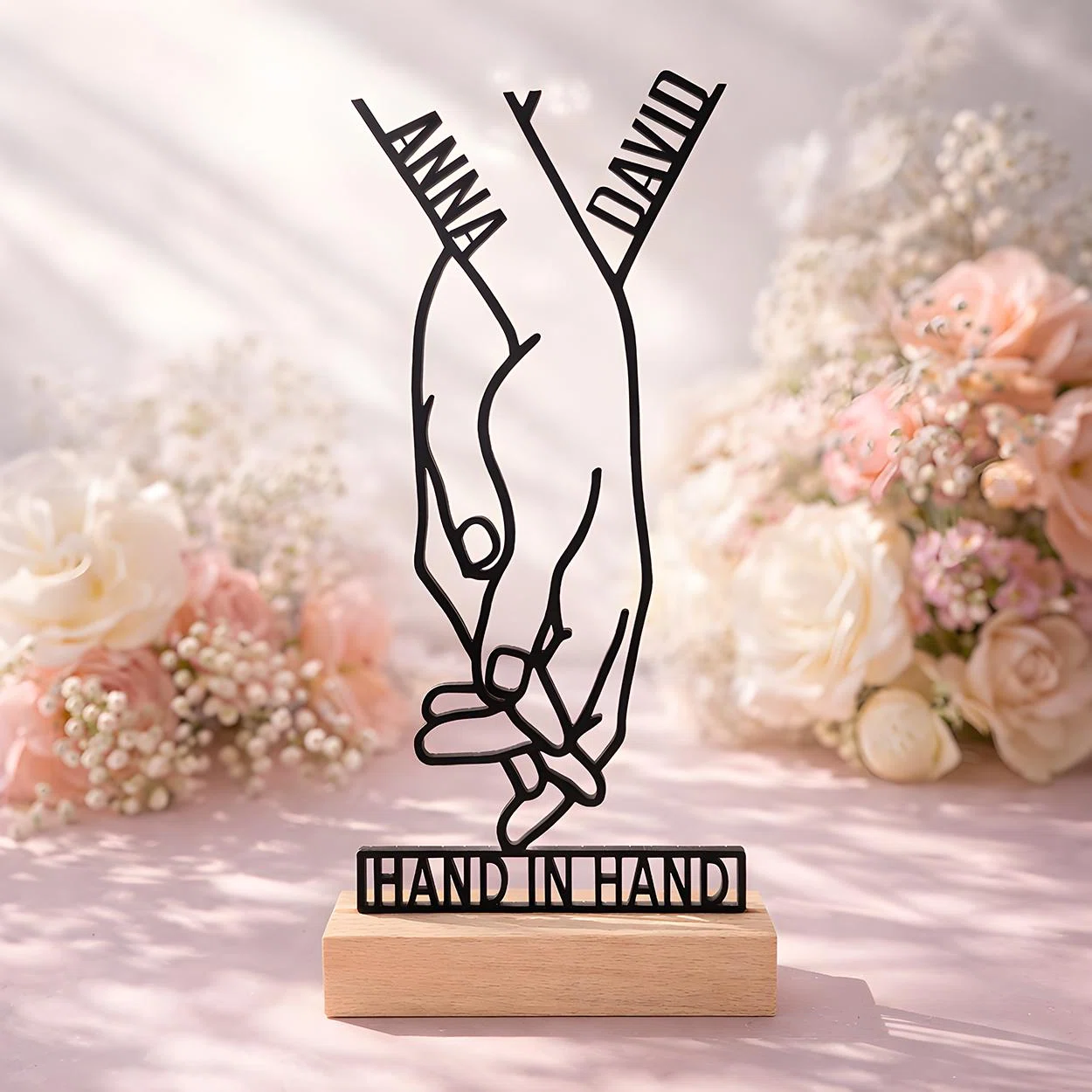 Hand-in-Hand-Aufsteller personalisiert bestellen: Zwei ineinandergreifende Hände bilden eine schwarze Linienfigur mit Schriftzug auf hellem Sockel, vor rosa-weißen Blumen.