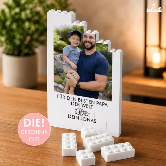 Personalisiertes Papa-Fotogeschenk bestellen: weißer Klemmbaustein-Aufsteller mit stufenförmigem Rand, Hochformatfoto, Papa-Widmung und Faustgruß-Motiv auf einem Holztisch.