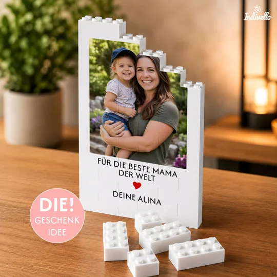 Fotogeschenk für Mama personalisiert bestellen: weißer Klemmbaustein-Aufsteller mit stufigem Rand, Foto, rotem Herz und der Widmung „Für die beste Mama der Welt“ auf einem Holztisch.