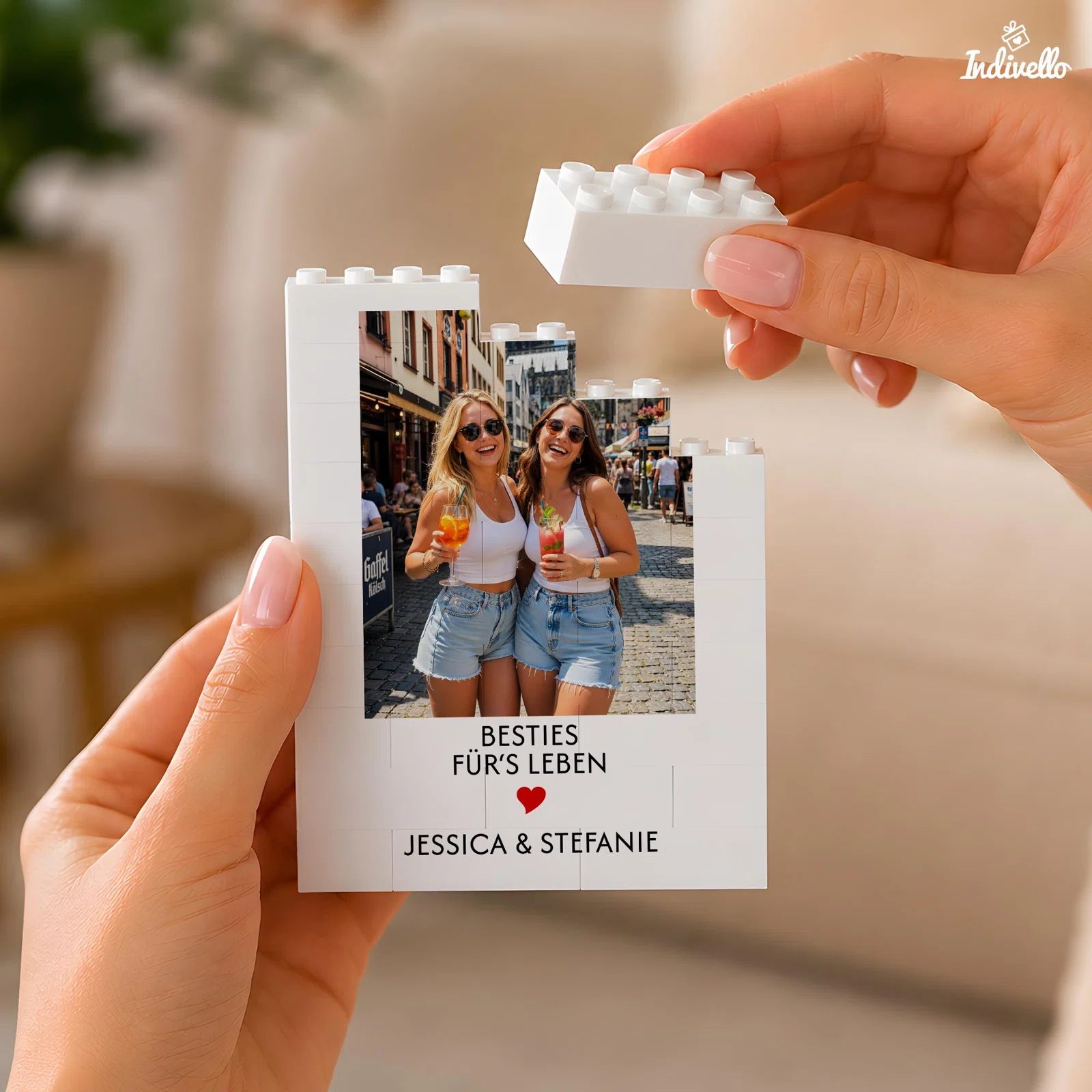 Weißes Fotomotiv aus Steckbausteinen, von zwei Händen zusammengesetzt. Es zeigt ein Urlaubsfoto zweier Personen sowie den Spruch „Besties für’s Leben“ und ein rotes Herz.