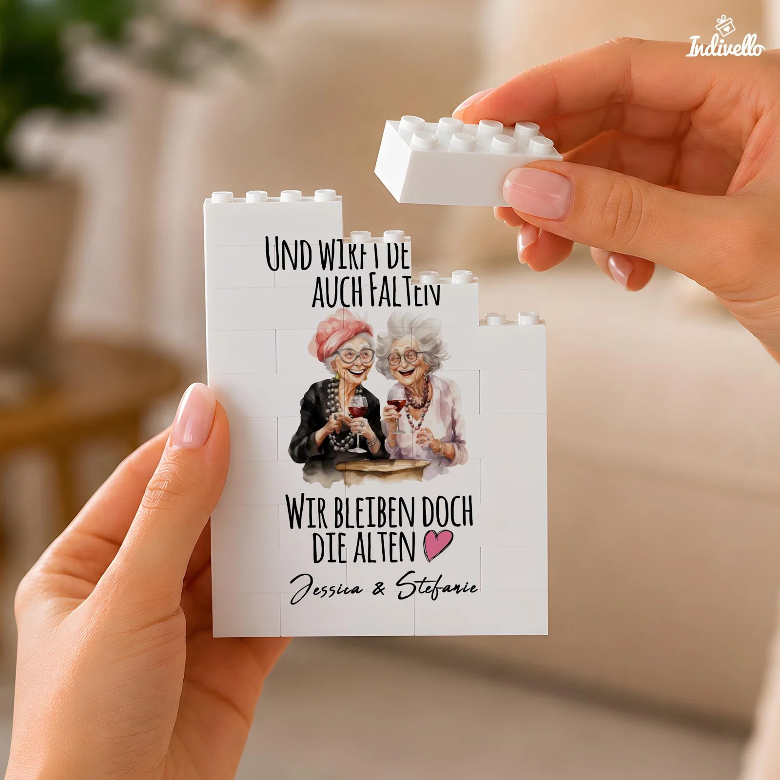 Personalisierbares weißes Klemmbaustein-Bild mit Illustration zweier älterer Frauen beim Weintrinken, rosa Herz und Spruch über Falten und „Wir bleiben doch die Alten“. Frontal in Händen gehalten.