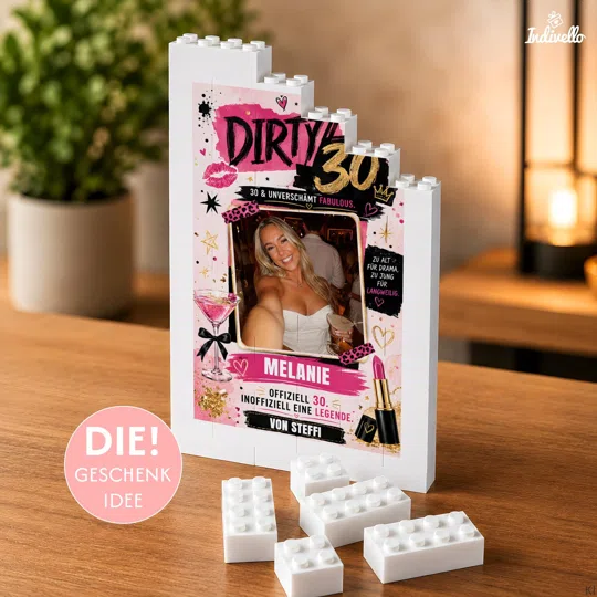 Personalisiertes Dirty-30-Fotogeschenk kaufen: weißer Klemmbaustein-Rahmen mit Partyfoto und pink-schwarz-goldenem Motiv, schräg von vorn auf einem Holztisch fotografiert.