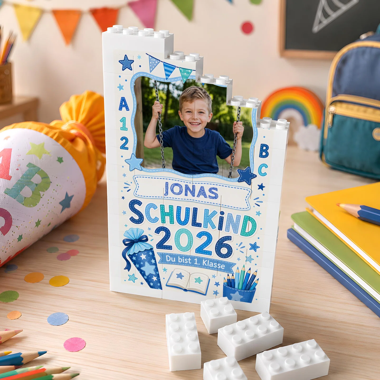 Weißer, treppenförmiger Fotorahmen im Baustein-Look, frontal auf einem Schreibtisch. Blaue und türkise Schulmotive, ABC, Zahlen sowie „Schulkind 2026“ umrahmen das Foto eines Kindes auf einer Schaukel.
