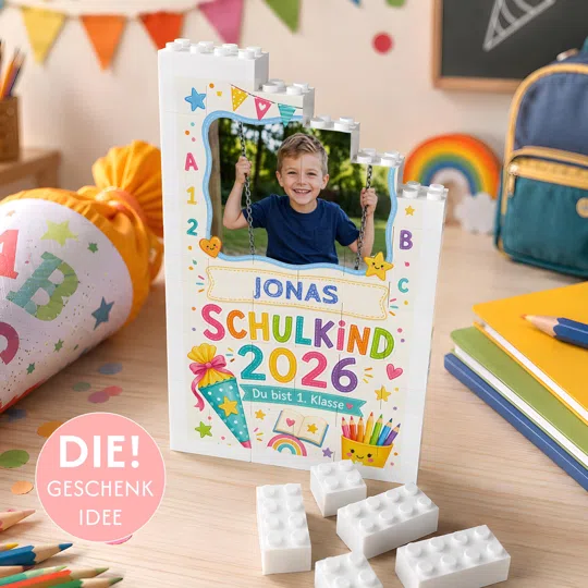Fotogeschenk zur Einschulung bestellen: weißer Baustein-Aufsteller mit Kinderfoto, bunten Schulmotiven, Name sowie „Schulkind 2026“ und „Du bist 1. Klasse“.