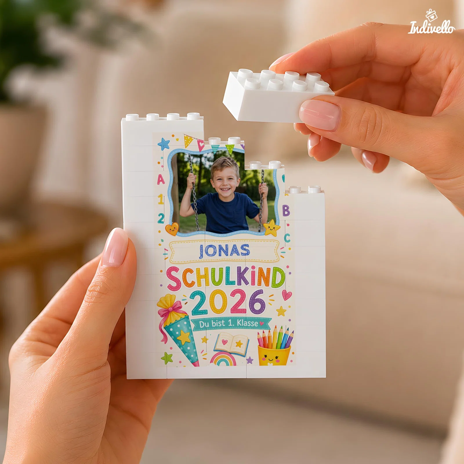 Personalisierter Fotobausatz aus weißen Klemmbausteinen zur Einschulung, mit buntem Schulkind-2026-Motiv, Namensfeld und Foto. Eine Hand setzt von oben einen Baustein auf.