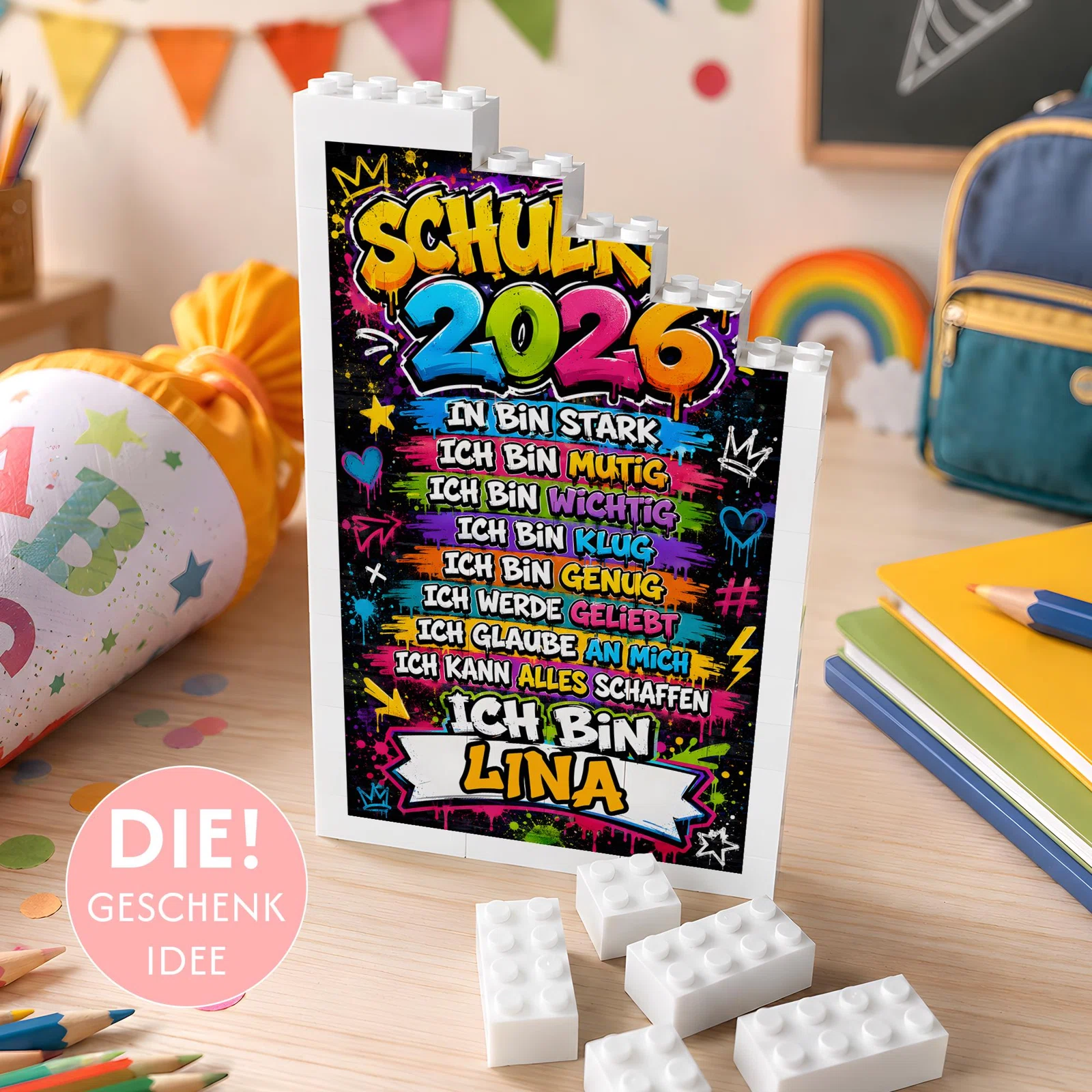 Personalisiertes Einschulungsgeschenk bestellen: weißer Baustein-Aufsteller mit bunter Graffiti-Schrift, stärkenden Ich-Botschaften, Jahreszahl 2026 und Namenszeile auf einem Schreibtisch.