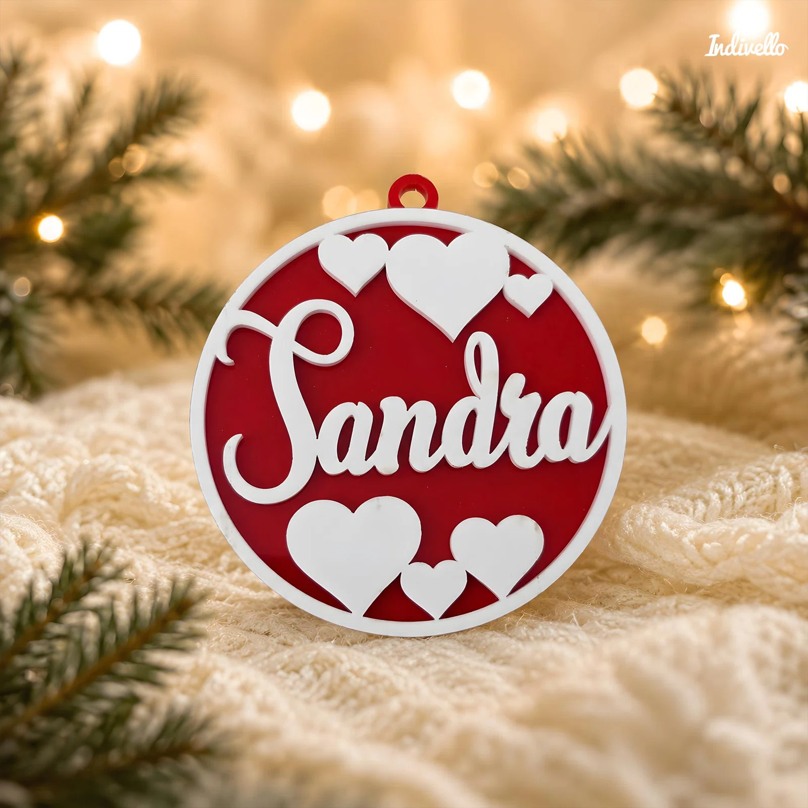 Runder, rot-weißer Weihnachtsanhänger mit dem geschwungenen Schriftzug „Sandra“, mehreren Herzmotiven und kleiner Öse. Frontal auf hellem Strick vor Tannenzweigen und warmen Lichtern arrangiert.