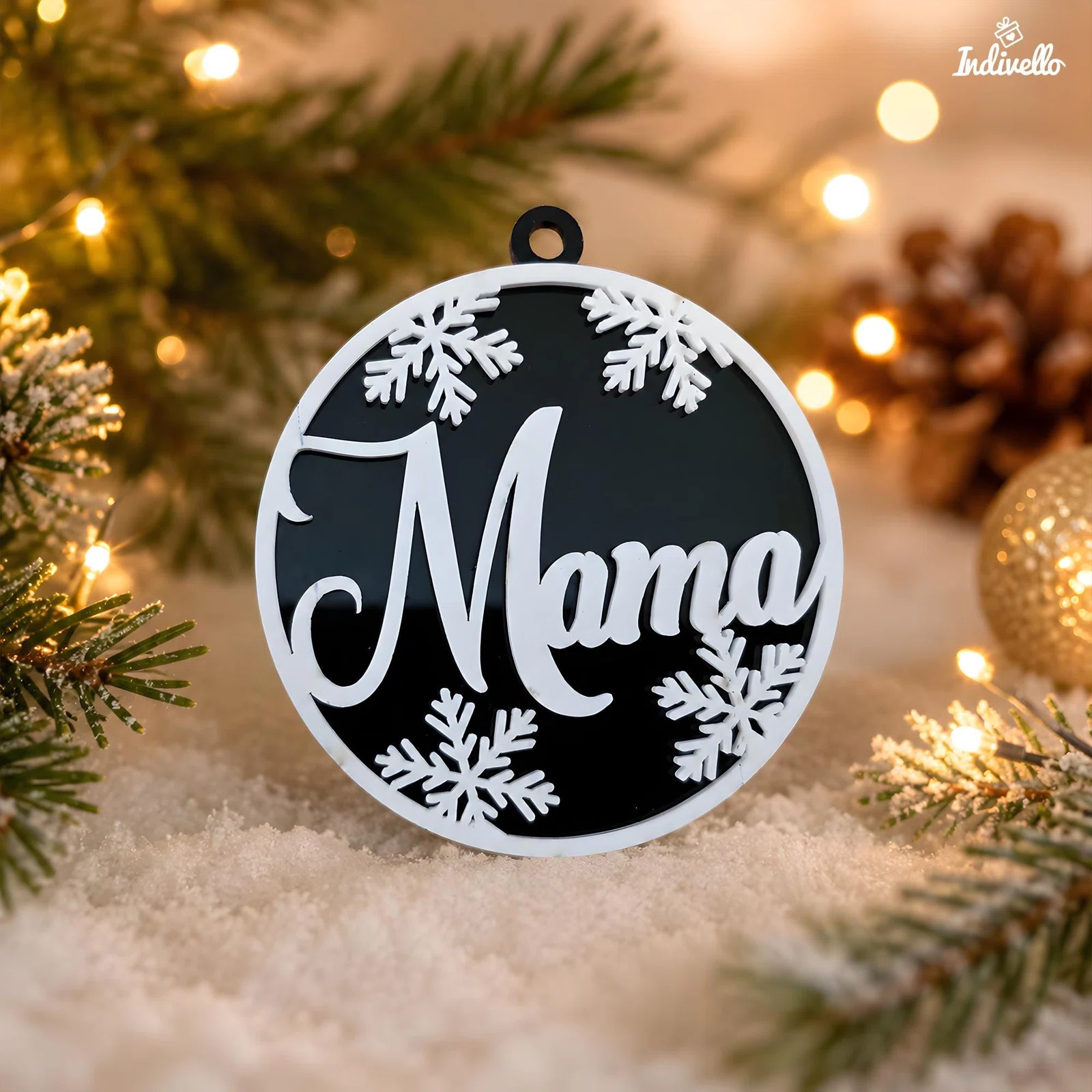 Runder schwarz-weißer Weihnachtsanhänger mit großer „Mama“-Schrift, Schneeflocken und Öse zum Aufhängen, frontal zwischen Tannenzweigen, Kunstschnee und warmen Lichtern dekoriert.