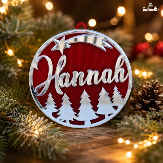 Weihnachtsanhänger mit Namen bestellen: runder, roter Anhänger mit silberfarbener Spiegelschrift, Sternschnuppe und Tannenwald, zwischen Tannenzweigen und warmen Lichtern.