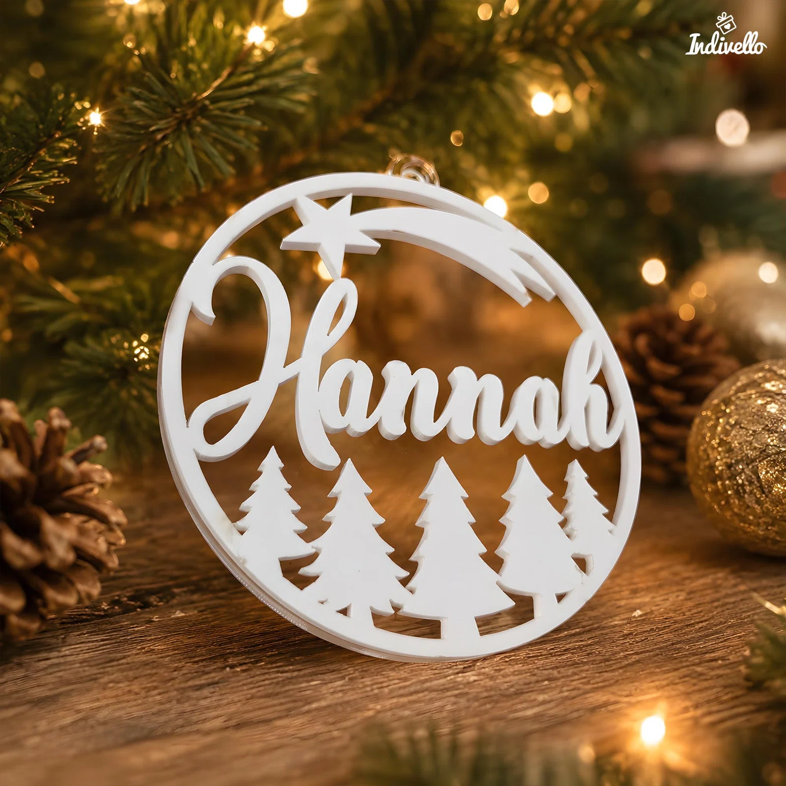 Runder weißer Weihnachtsanhänger mit dem Schriftzug „Hannah“, Sternschnuppe und ausgeschnittenen Tannenbäumen. Er steht vor einem beleuchteten Weihnachtsbaum auf einer Holzfläche.