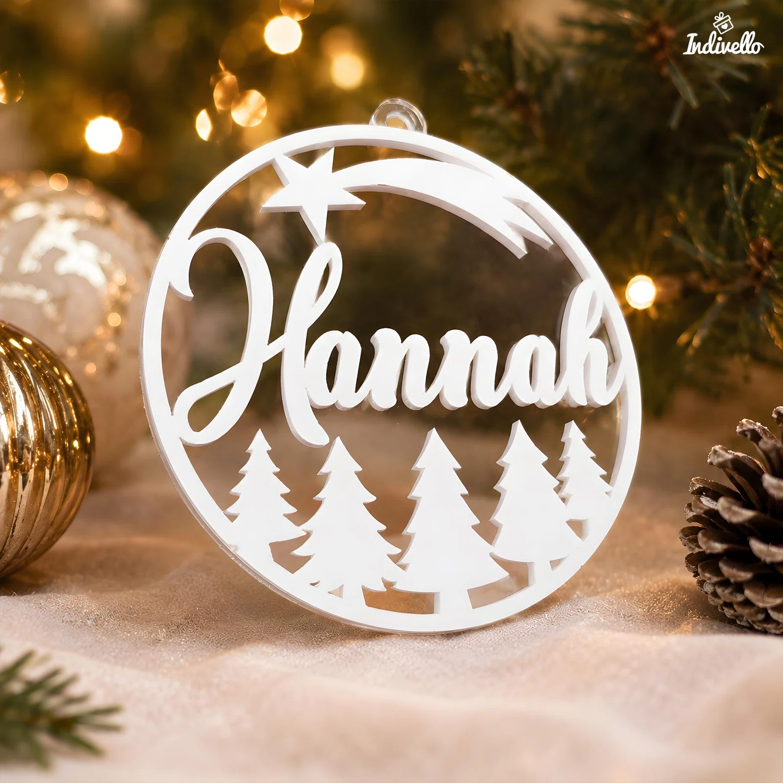 Runder, weißer Weihnachtsanhänger mit dem Schriftzug „Hannah“, Sternschnuppe und Tannenwald. Frontal zwischen warmen Lichtern, Christbaumzweigen, Kugeln und einem Tannenzapfen inszeniert.