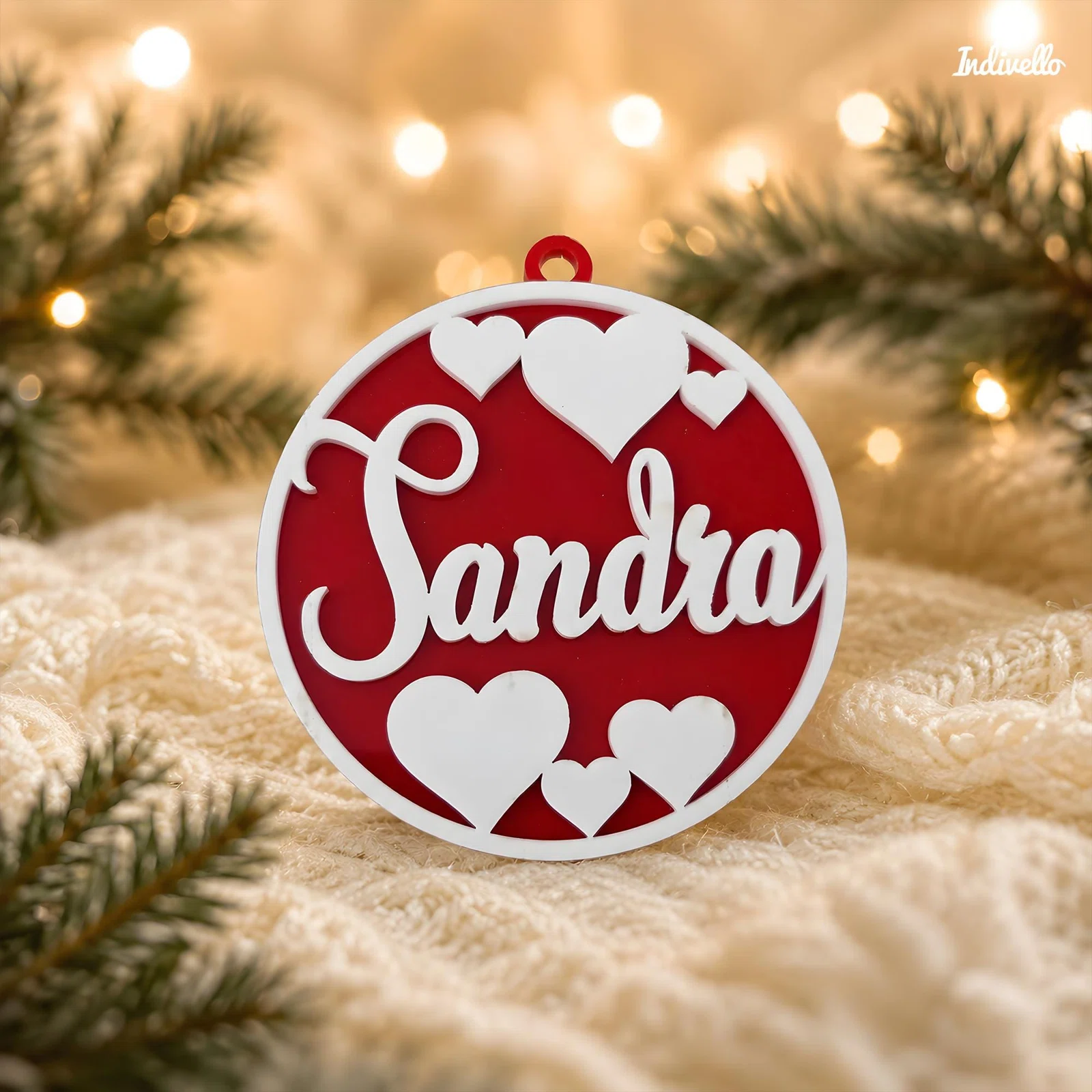 Runder rot-weißer Weihnachtsanhänger mit dem Schriftzug „Sandra“, mehreren Herzmotiven und kleiner Öse, frontal zwischen Tannenzweigen und warmen Lichtern inszeniert.