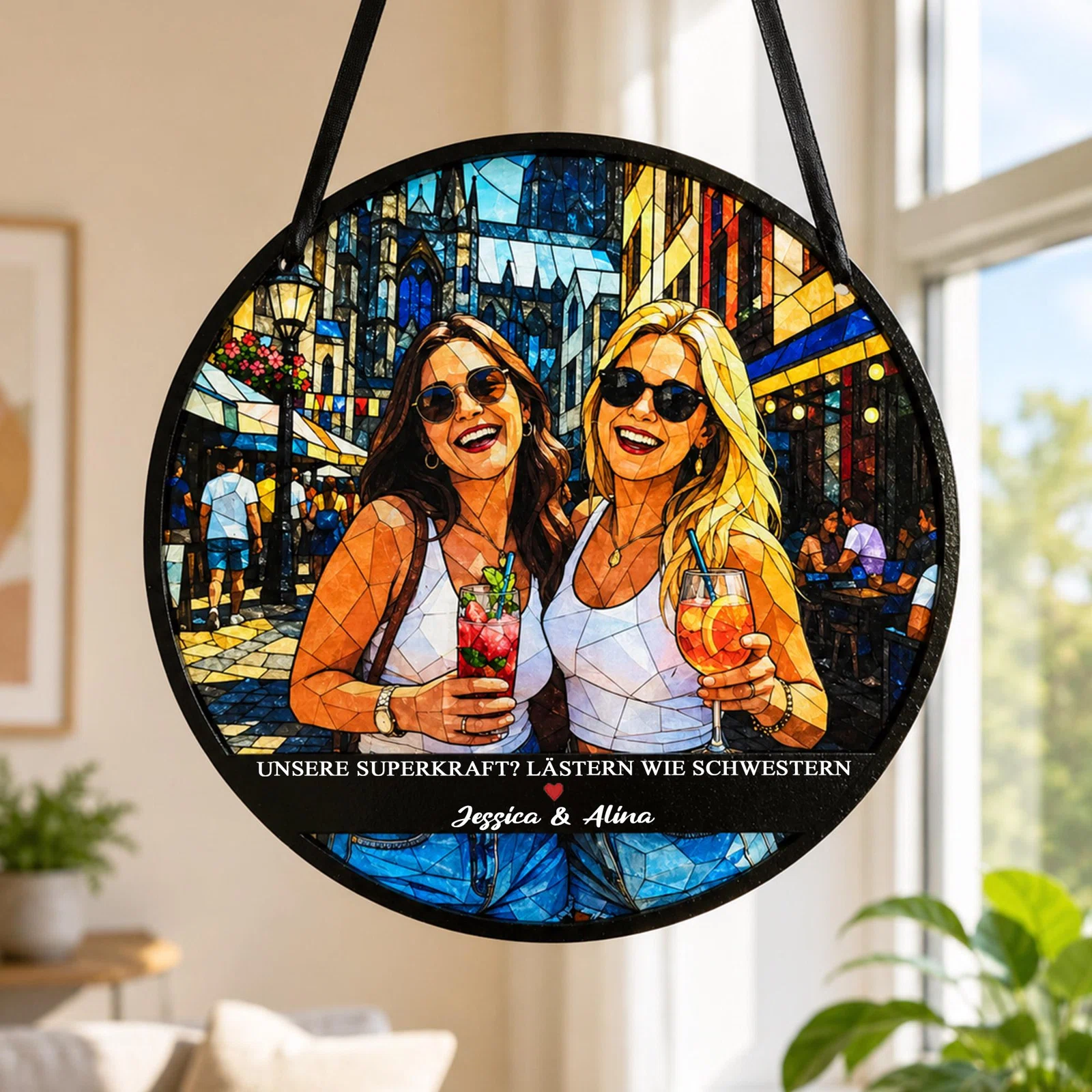 Runder Fensteranhänger im farbenfrohen Buntglas-Look mit zwei Frauen in Sonnenbrillen und Getränken vor einer Stadtkulisse. Darunter steht der Spruch „Unsere Superkraft? Lästern wie Schwestern“ samt Namen.