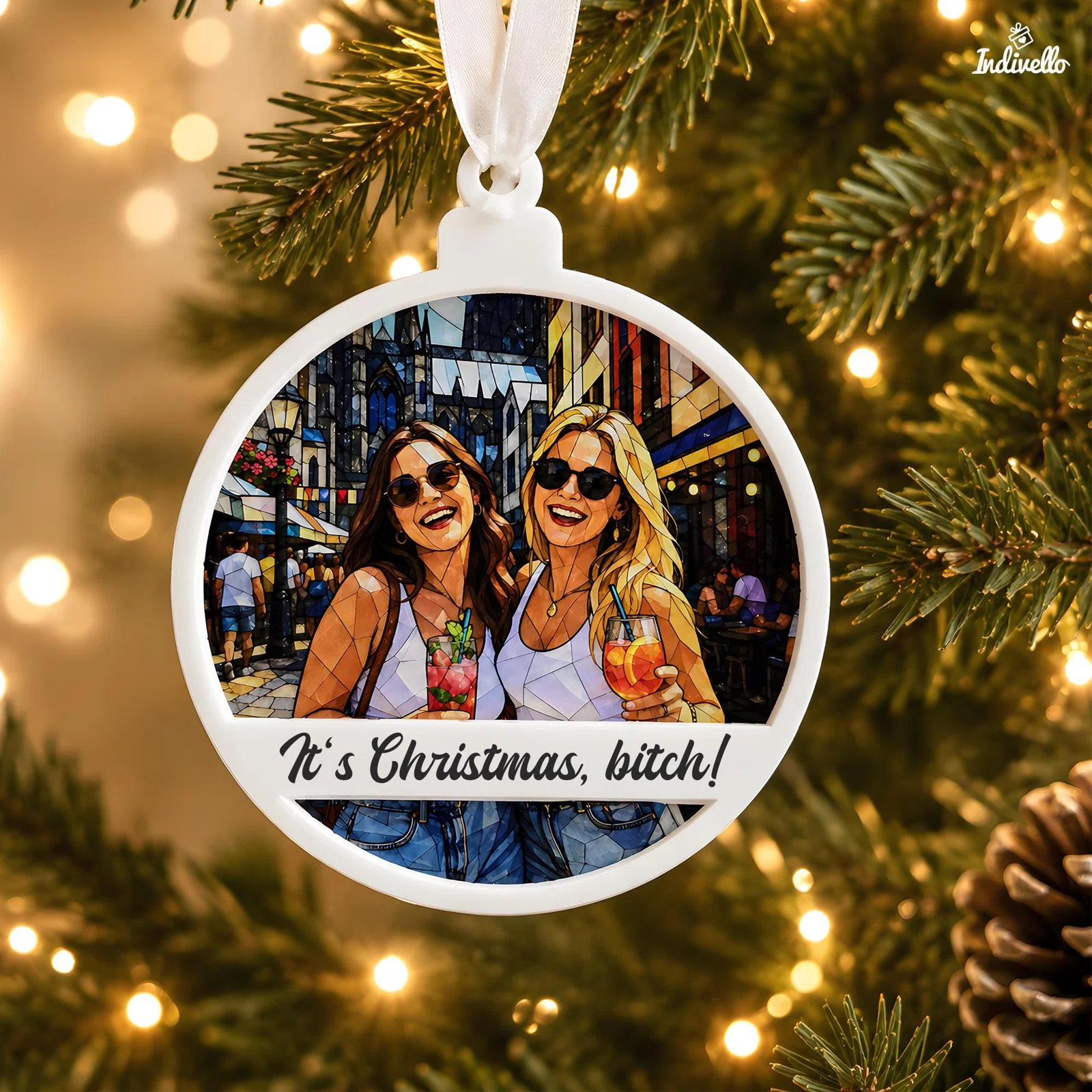 Runder, weiß gerahmter Weihnachtsanhänger mit farbigem Mosaikmotiv zweier Frauen mit Sonnenbrillen und Getränken sowie dem Schriftzug „It’s Christmas, bitch!“, nah am beleuchteten Tannenbaum.
