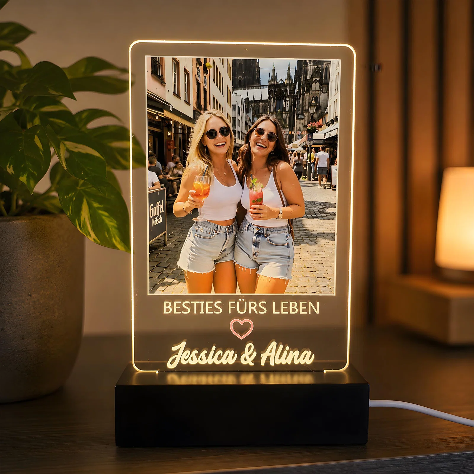 Rechteckige Fotoleuchte mit warmweiß leuchtendem Rand auf schwarzem Sockel. Das Stadtfoto zweier Personen mit Getränken wird durch „Besties fürs Leben“, ein Herz und zwei Namen ergänzt.