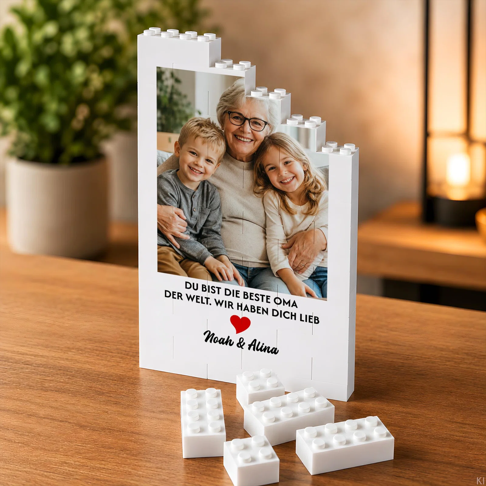 Weißer, aufrecht stehender Fotorahmen in Baustein-Optik mit abgestufter Noppenkante, Foto dreier Personen und der Widmung „Du bist die beste Oma der Welt, wir haben dich lieb“ samt rotem Herz.