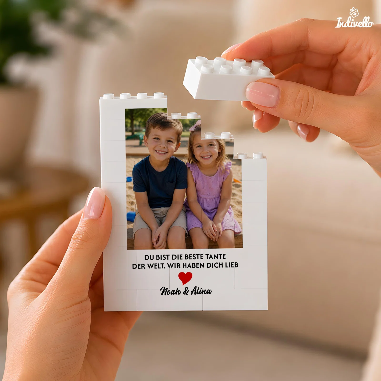 Personalisierbarer weißer Bilderrahmen in Klemmbaustein-Optik, frontal in Händen gehalten. Unter dem Kinderfoto stehen ein Tante-Spruch, ein rotes Herz und zwei Namen.