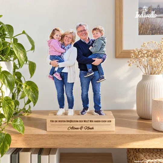 Freistehender Fotoaufsteller mit ausgeschnittener Ganzkörperaufnahme von zwei lächelnden Erwachsenen, die jeweils ein Kind auf dem Arm halten. Auf dem rechteckigen Sockel steht „Für die besten Großeltern der Welt! Wir haben euch lieb – Alina & Noah“; frontal auf einem Holztisch zwischen grüner Pflanze, Vase und Kerze präsentiert.