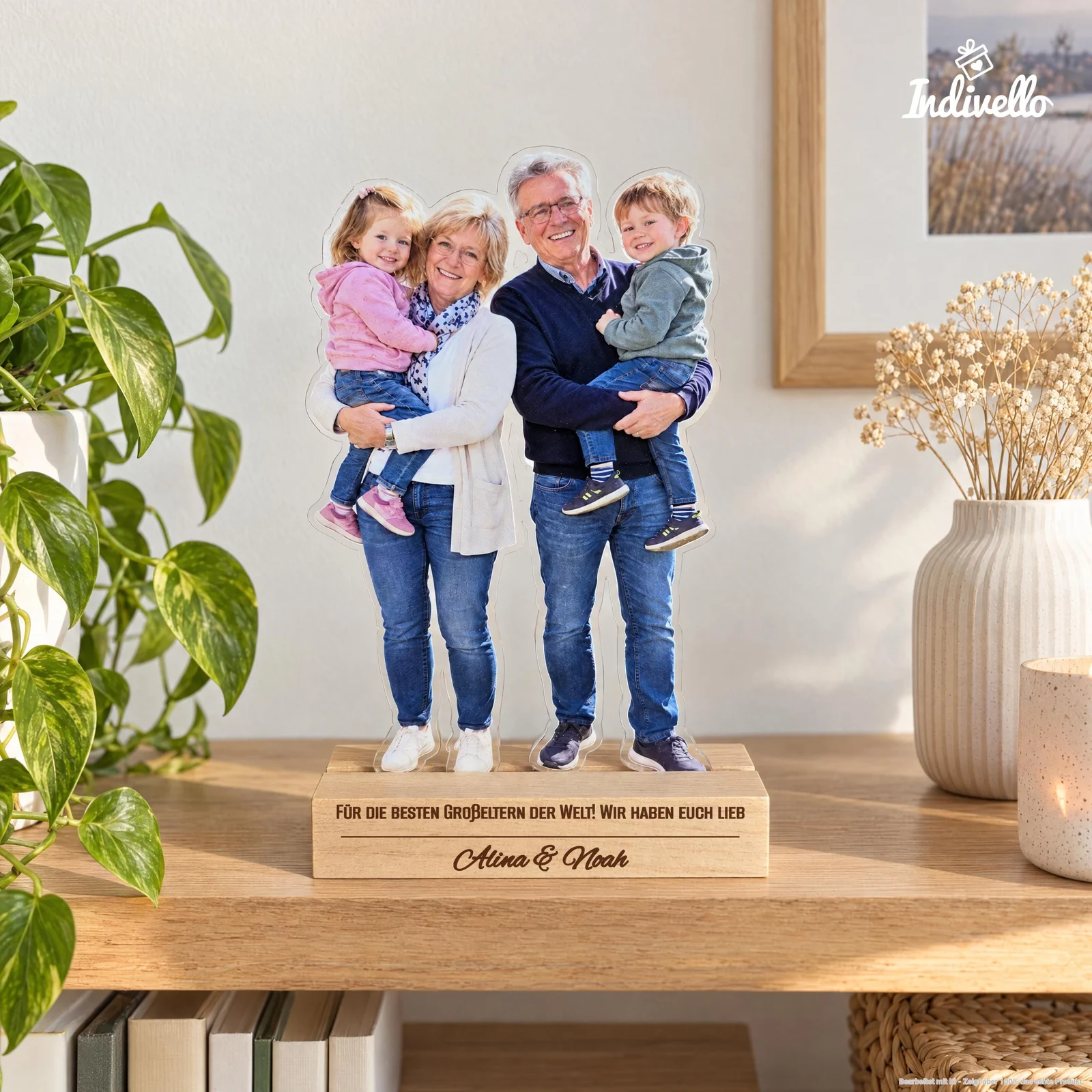 Freistehender Fotoaufsteller mit ausgeschnittener Ganzkörperaufnahme von zwei lächelnden Erwachsenen, die jeweils ein Kind auf dem Arm halten. Auf dem rechteckigen Sockel steht „Für die besten Großeltern der Welt! Wir haben euch lieb – Alina & Noah“; frontal auf einem Holztisch zwischen grüner Pflanze, Vase und Kerze präsentiert.