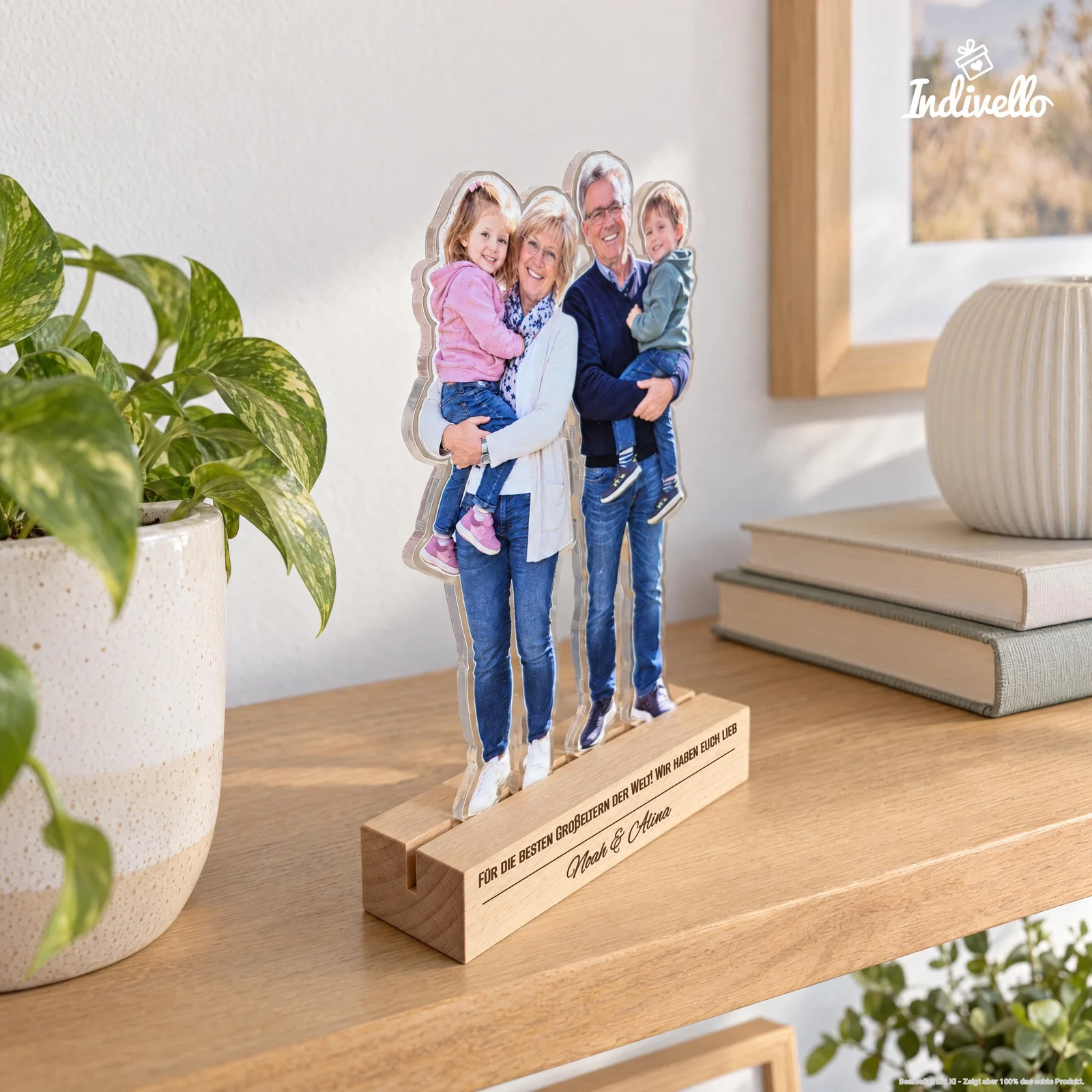 Frontal gezeigter, transparenter Fotoaufsteller mit der ausgeschnittenen Kontur von zwei Erwachsenen, die jeweils ein Kind im Arm halten. Er steht auf einem hellen Sockel mit der Aufschrift „Für die besten Großeltern der Welt. Wir haben euch lieb – Noah & Alina“ auf einem Holzregal zwischen grüner Pflanze, Büchern und Vase.