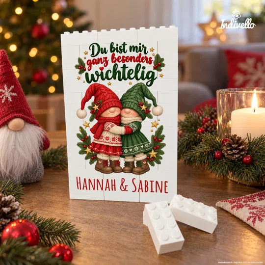 Frontansicht eines weißen Klemmbaustein-Bildes mit zwei sich umarmenden Weihnachtswichteln in Rot und Grün, Tannenzweigen und dem Spruch „Du bist mir ganz besonders wichtelig“ sowie den Namen „Hannah & Sabine“. Es steht auf einem festlich dekorierten Holztisch neben Kerze, Zweigen, roten Kugeln und einzelnen weißen Bausteinen.