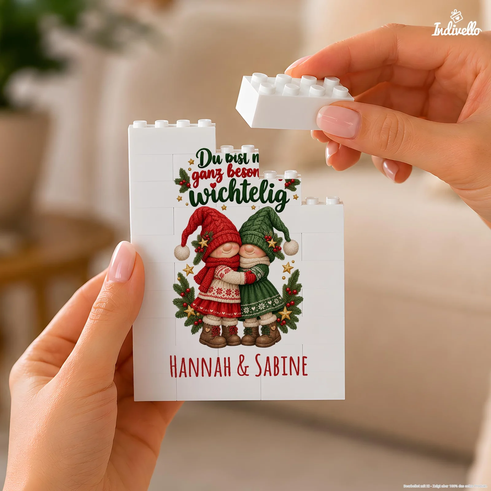 Nahaufnahme einer weißen Klemmbaustein-Platte, die von zwei Händen gehalten und oben mit einem Stein ergänzt wird. Das weihnachtliche Motiv zeigt zwei sich umarmende Wichtel in Rot und Grün, Tannenzweige und Sterne sowie den Text „Du bist mir ganz besonders wichtelig“ und „Hannah & Sabine“.
