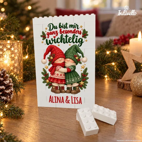 Frontansicht einer weißen, aus Klemmbausteinen aufgebauten Bildtafel mit zwei sich umarmenden Weihnachtswichteln in Rot und Grün. Darüber steht „Du bist mir ganz besonders wichtelig“, darunter „ALINA & LISA“; die Tafel steht zwischen Tannenzweigen, Lichtern und Kerzen auf einem Holztisch.